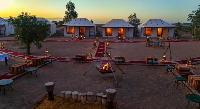 Merzouga desert camp at dusk: white tents with lit windows, lantern-lined carpet paths and a fire pit